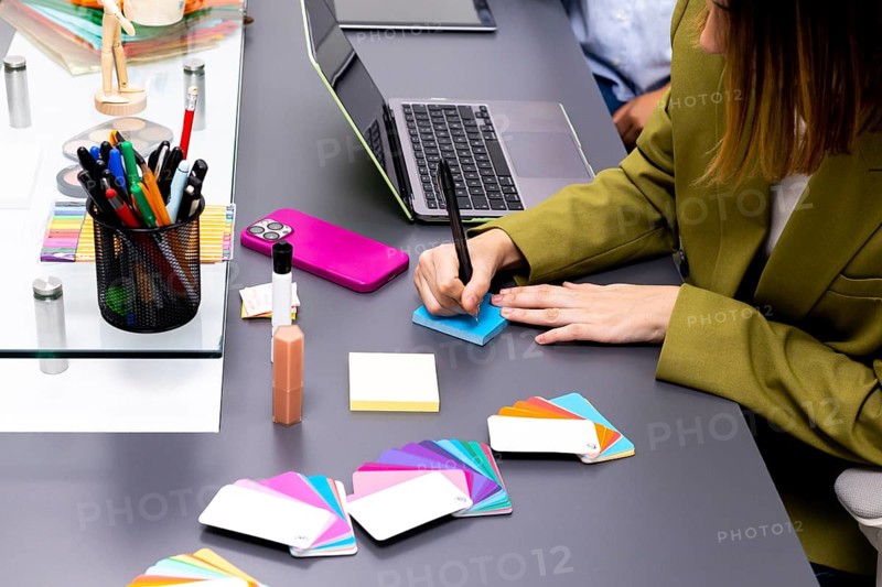 A professional consultant in an office uses color swatches and notes to develop branding strategies. The scene includes a laptop, colorful pens, and a phone for creative work