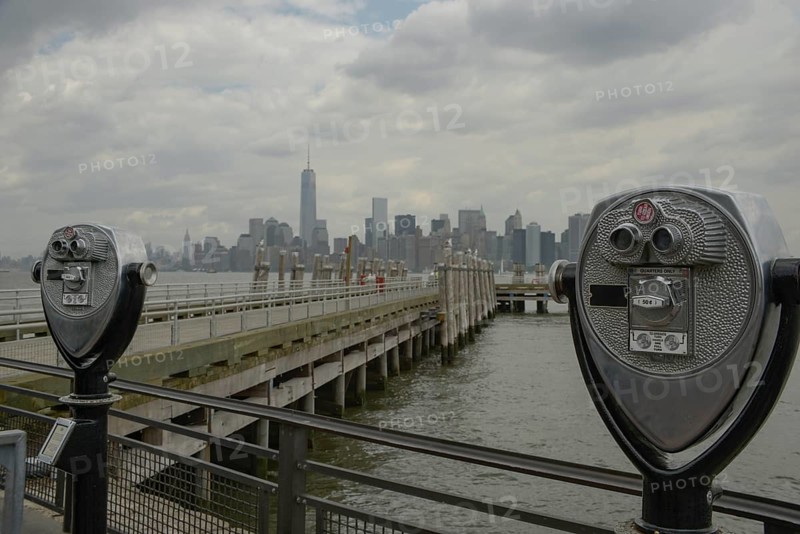 A scenic view of Manhattan from a pier at Liberty State Park, featuring a coin-operated binoculars foregrounding the iconic skyline
