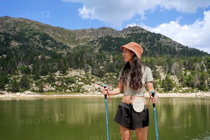 A woman with an ostomy bag stands confidently by a serene mountain lake, wearing a hat and holding trekking poles The scene reflects empowerment and adventure