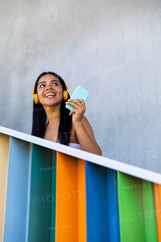 A joyful woman with headphones smiles while using her smartphone against a backdrop of vibrant modern architecture, conveying a sense of connection and technology