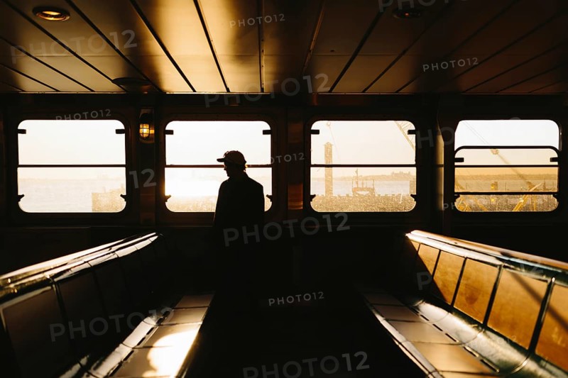 A man in silhouette stands inside a ferry, gazing out at a serene, sunlit harbor scene in New York. The warm, golden sunlight casts an atmospheric glow across the interior