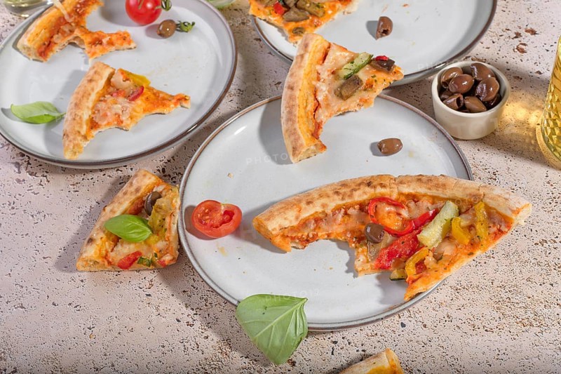 Slices of vegetarian pizza with basil, cheese, and mixed vegetables sit invitingly on plates. The scene captures a casual, delicious snack setting with a refreshing drink