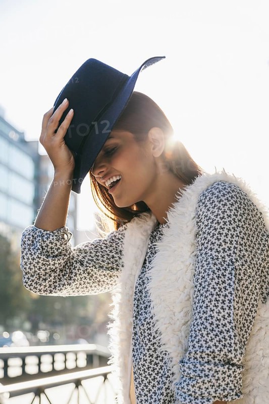 A woman in a patterned shirt and furry vest joyfully holds her hat, enjoying the sunlight. Her relaxed pose and smile enhance the vibrant, urban setting behind her