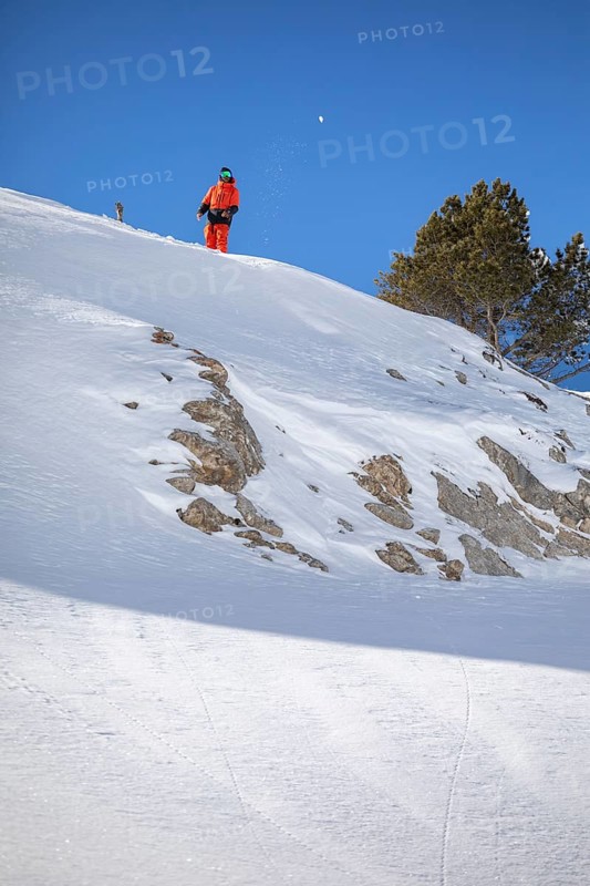 A snowboarder in orange gear stands on a snow covered mountain slope. The bright blue sky contrasts with the pristine snow, capturing a serene winter scene
