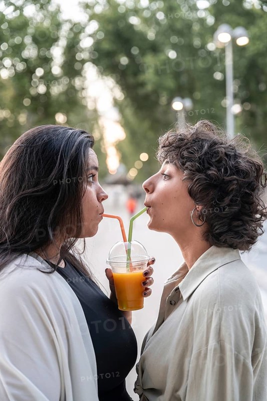 A lesbian couple shares a juice through straws in a tender moment, standing outdoors with a blurred, leafy background. The scene captures love and connection