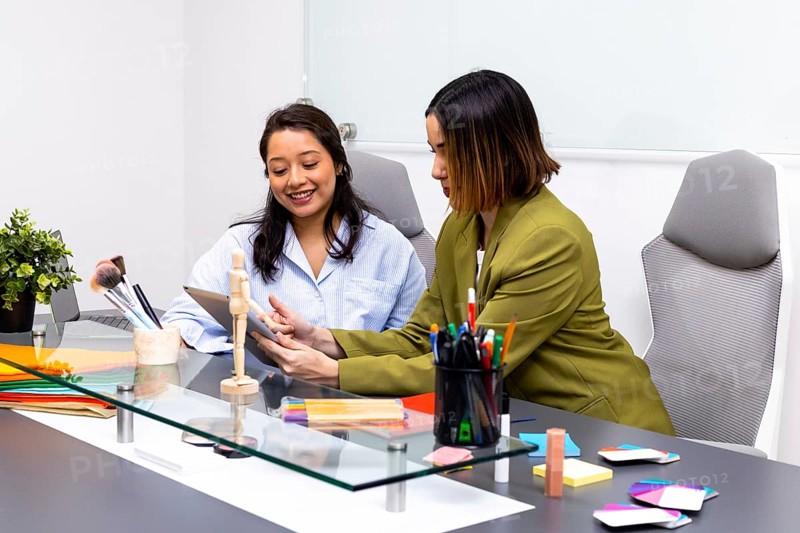 Two women engage in an image consulting session in a modern office. One shows color swatches while the other holds a tablet, surrounded by pens, brushes, and papers