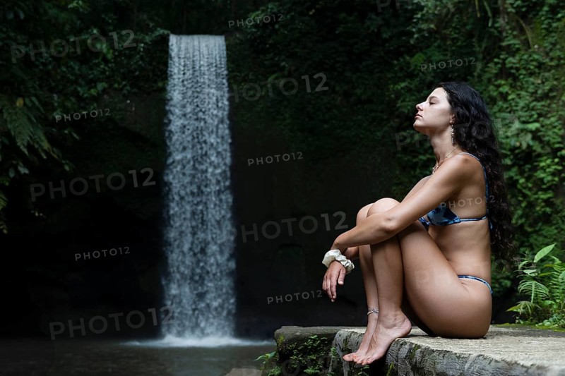 A woman sits peacefully in meditation, surrounded by lush greenery and a cascading waterfall in Bali. Her relaxation and connection to nature create a tranquil scene