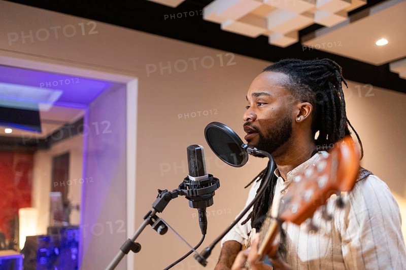 A black musician passionately sings into a microphone while playing guitar in a professional recording studio, with acoustic panels and modern equipment enhancing the ambiance