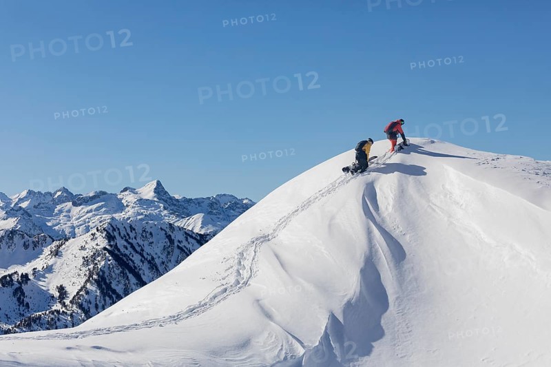 Two snowboarder friends make their way up a pristine snow covered peak, leaving tracks behind. The majestic mountain backdrop complements the clear blue sky, embodying adventure