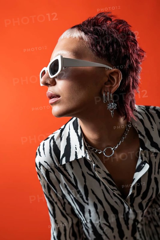 Fashion model wearing animal print shirt, white sunglasses, and silver accessories in front of a bright orange backdrop. Studio setting with flash photography accentuates style