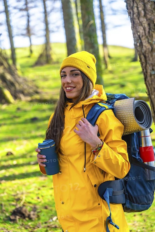 Young hiker woman wearing a yellow raincoat and backpack is holding a hot drink in a forest during a trekking day