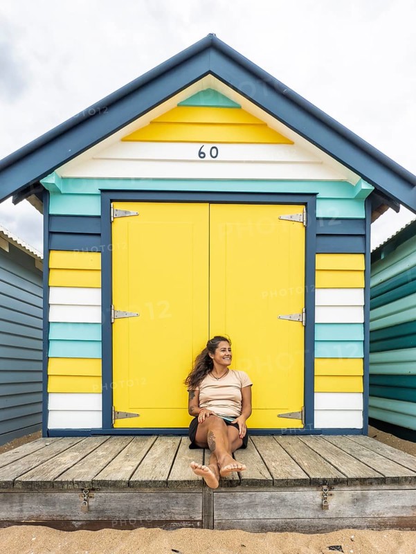 A cheerful scene of the iconic Brighton Bathing Boxes in Melbourne, Australia, showcasing vibrant colors with a woman sitting relaxed in front of a yellow door