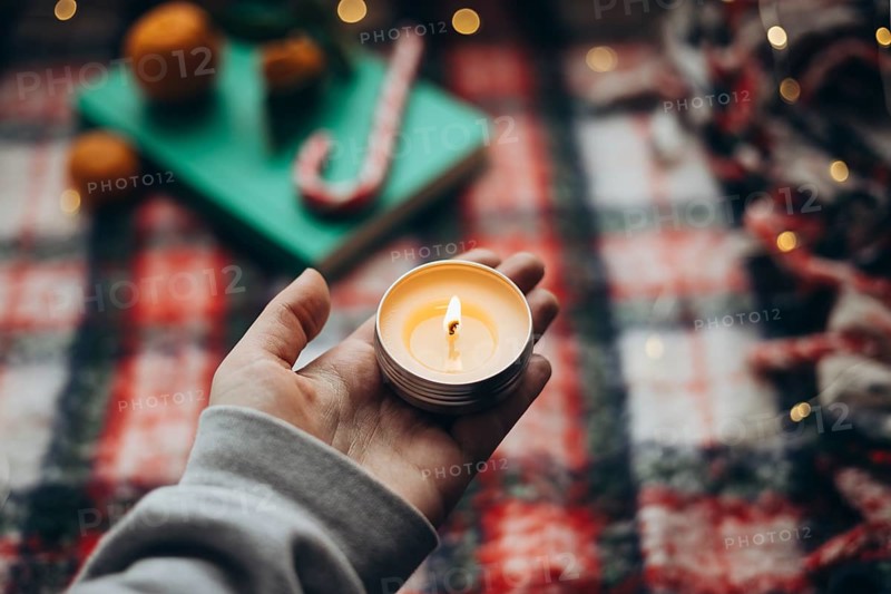 Cropped unrecognizable a person's hand holding a lit candle, with a blurred festive backdrop showing hints of oranges and candy canes, embodying the warmth and cheer of the Christmas season