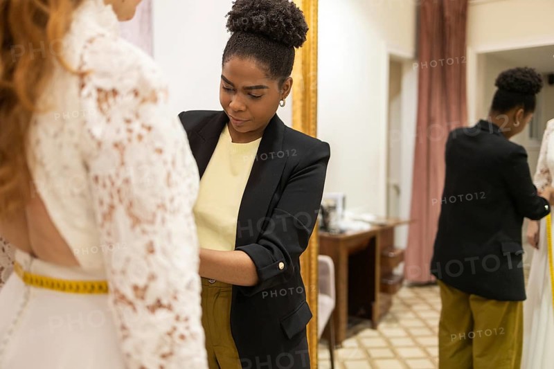 A seamstress skillfully adjusts a lace dress on a client in a tailor studio. The setting is elegant, with a large mirror reflecting the attention to detail