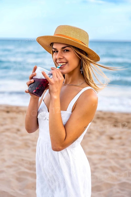 Young woman wearing straw hat and white dress drinking refreshing beverage on beach during summer vacation