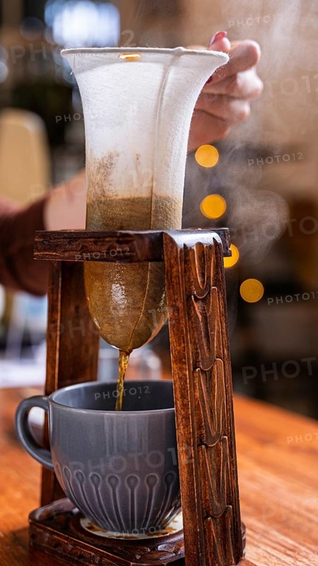 A cozy cafe scene showcasing a traditional coffee brewing method with a cloth filter. The steaming beverage pours into a ceramic mug, evoking warmth and simplicity