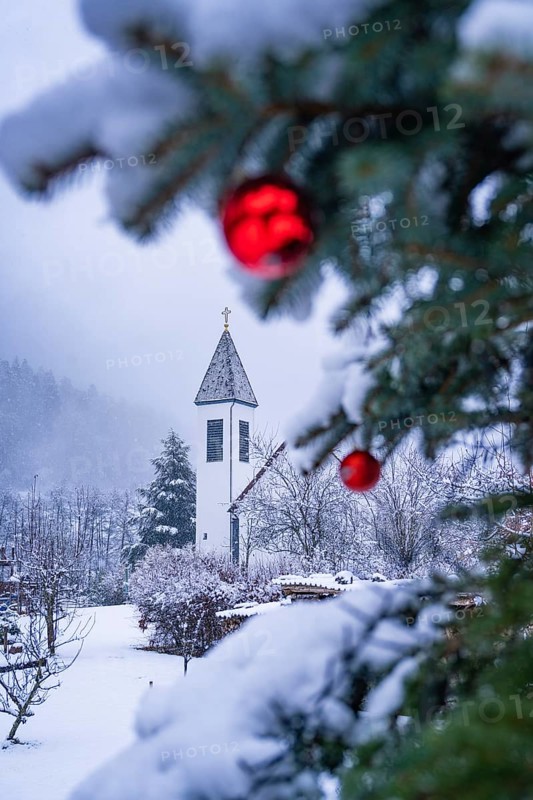 Focus on red balls and snowy branches in front of a church tower in the background, Enzklösterle, district of Calw, Black Forest, Germany