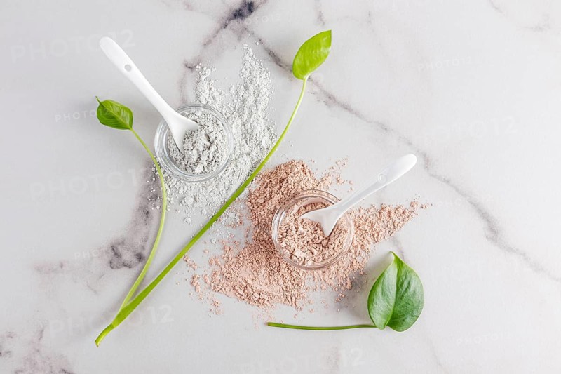 Top view of natural cosmetic clay powders in clear bowls with spoons on a marble surface, accompanied by fresh green leaves, used for skincare and beauty treatments