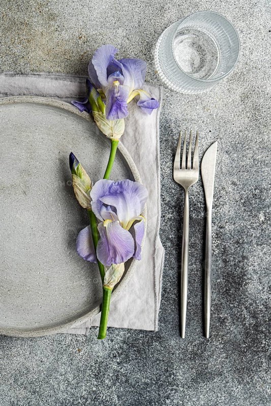From above, an elegant table setting features a purple iris flower alongside a gray ceramic plate, a glass of water, and silver cutlery on a textured gray background