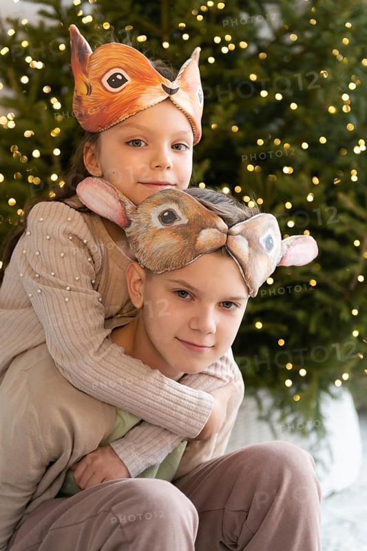 A girl and boy embrace, wearing animal masks, with a beautifully lit Christmas tree in the background