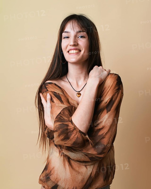 Portrait of a young woman on a beige background, captured in a studio setting. She is smiling and dressed in a brown patterned blouse, expressing a joyful demeanor