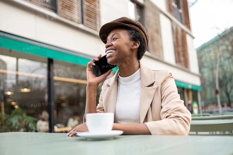 A smiling black woman sits at an outdoor cafe, enjoying a phone conversation She radiates happiness, embodying the vibrancy of urban life amidst a modern city backdrop