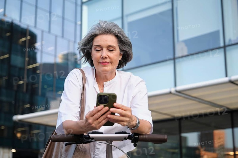 Stylish mature woman checking her smartphone while leaning on a scooter in a modern urban environment. She looks engaged and tech-savvy in casual attire