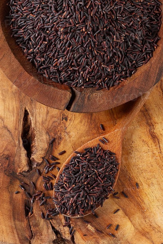 Natural black rice in a wooden bowl and spoon on a wooden surface in rustic style, top view