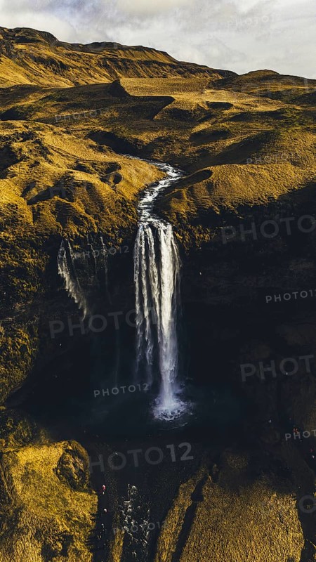 A stunning aerial view of Seljalandsfoss waterfall in Iceland, with water cascading down a high cliff, surrounded by golden, mossy terrain and winding river paths
