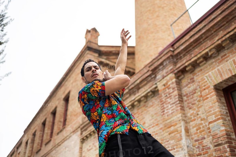 A Cuban man, dressed in a colorful shirt, passionately dances against a historic brick building backdrop. His dynamic pose captures the essence of cultural expression and rhythm