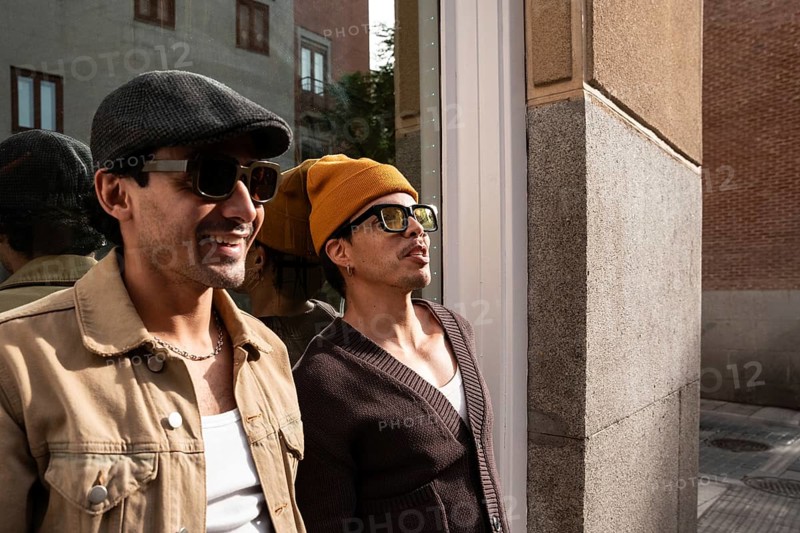 A Colombian gay couple smiles while enjoying a sunny day in the city. Both are wearing stylish sunglasses and casual attire, exuding happiness and confidence