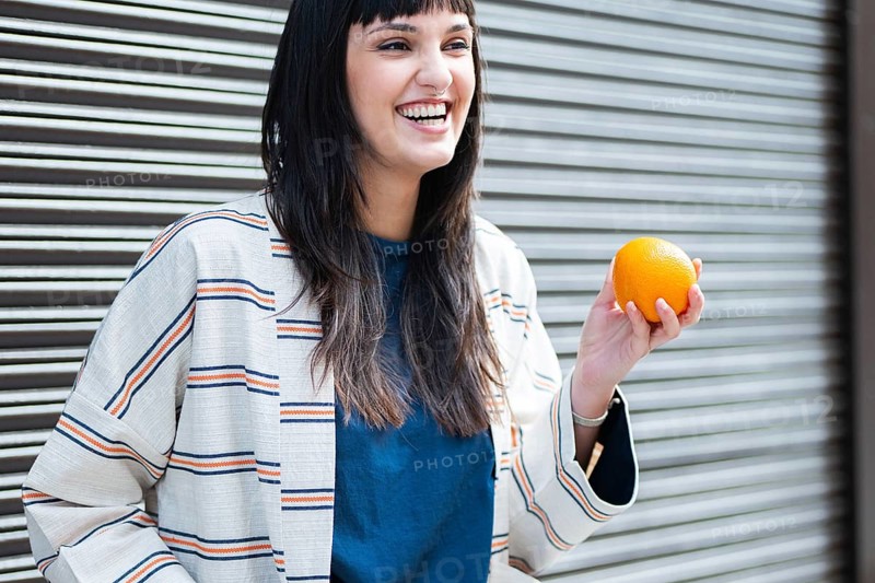 A cheerful woman with a striped jacket holds a bright orange, smiling broadly against a metallic shutter. Her joy and casual attire create a vibrant and lively scene