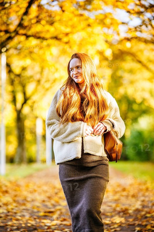 Beautiful pregnant woman with long red hair enjoys a serene walk through a sunlit autumn park. She wears a warm cozy outfit, surrounded by vibrant golden foliage