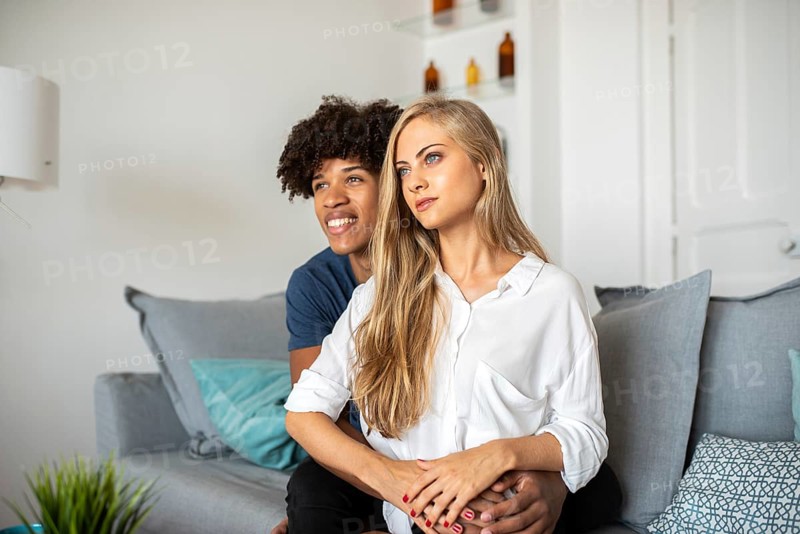 A couple sits closely on a comfortable sofa, sharing a peaceful moment. They appear relaxed and content, surrounded by soft pillows in a bright, welcoming room