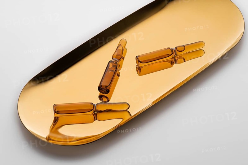 Three amber-colored cosmetic ampoules containing anti-aging serum, elegantly displayed on a reflective golden tray, ideal for skincare routines