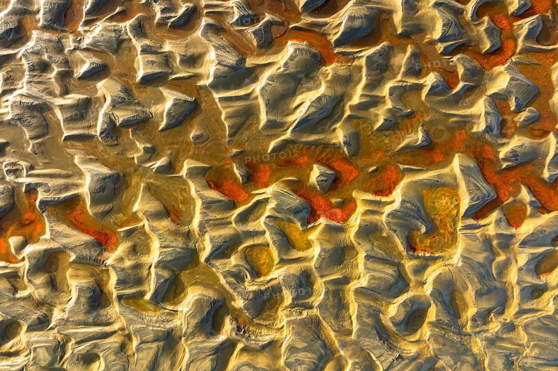 Aerial image of sand dunes showcasing intricate textures and patterns, highlighted by warm, golden sunlight. Rich red and orange hues add depth to the unique landscape