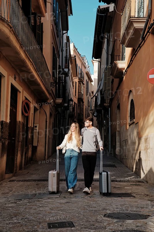 A young couple strolls through the charming streets of Majorca, Spain, their luggage in tow, seeking the ideal vacation home. The warm sun and historic architecture set a romantic scene