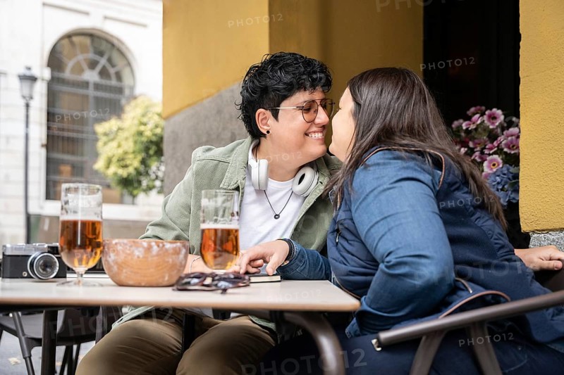 A joyful curvy lesbian couple shares a romantic moment at a bar, sitting close with smiles. Two glasses of beer and a camera sit on the table, enhancing the cozy atmosphere