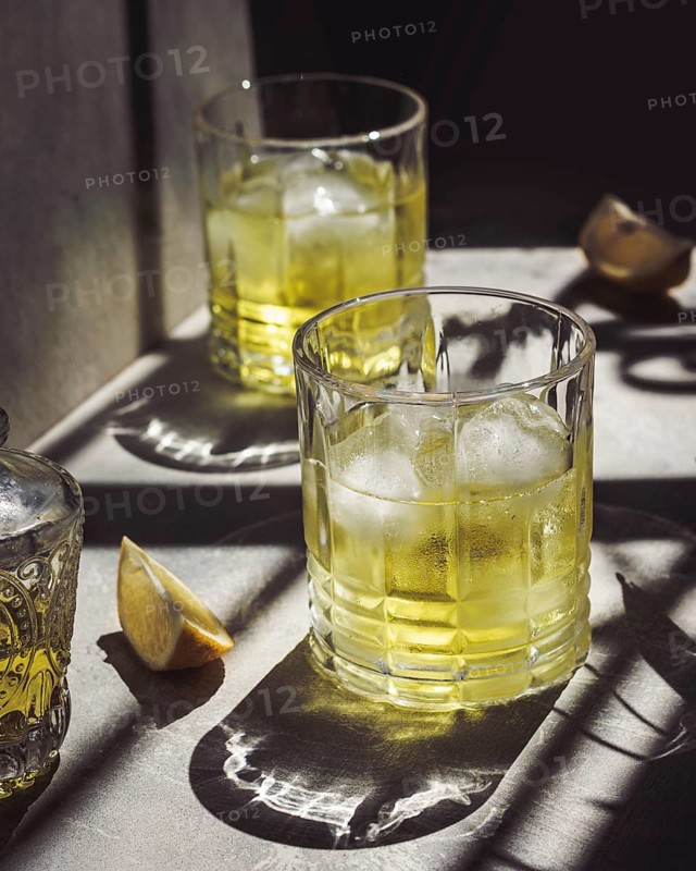 Glasses of refreshing lemon liqueur with ice cubes shine in the sunlight, casting intricate shadows. The scene evokes a vibrant and inviting atmosphere perfect for relaxation