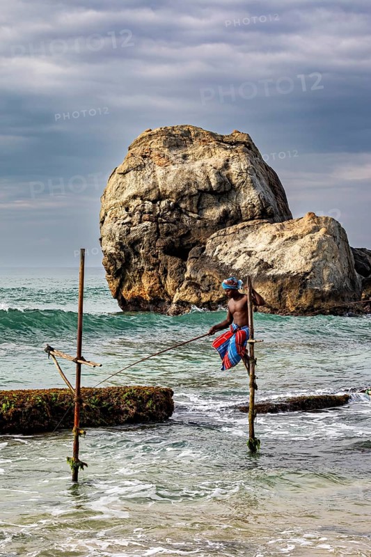 A fisherman sits on a wooden pole in the water, in front of a majestic rocky landscape, The stilt fishermen on the beach near Galle in Sri Lanka