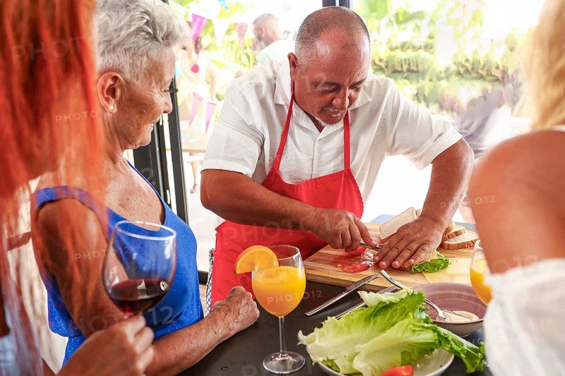 A man in a red apron slices tomatoes while chatting with senior women at an outdoor party, displaying a joyous atmosphere among friends over 50 enjoying a social gathering