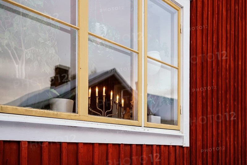 A warm window scene in Swedish Lapland. Yellow-framed window reflects a cozy, candle-lit interior with potted plants, set against the iconic red wooden exterior