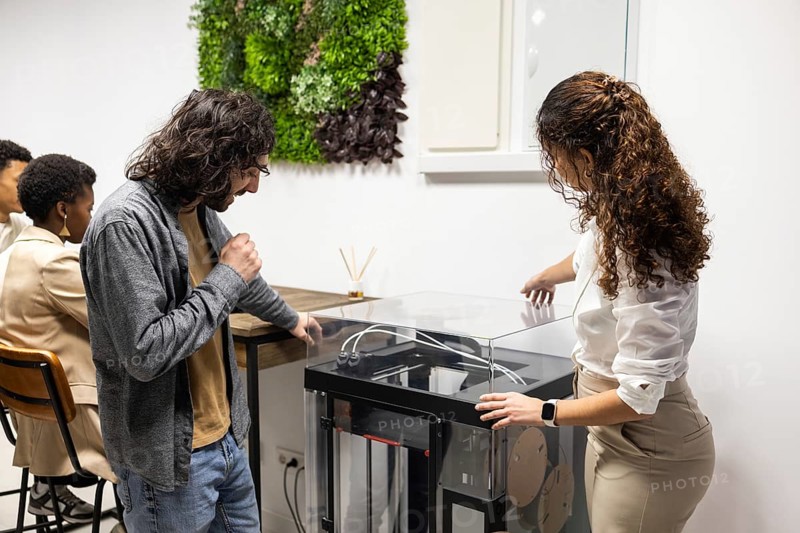 A diverse team collaborates in a modern office. Colleagues engage with a 3D printer while others focus on tasks, creating an atmosphere of innovation and teamwork