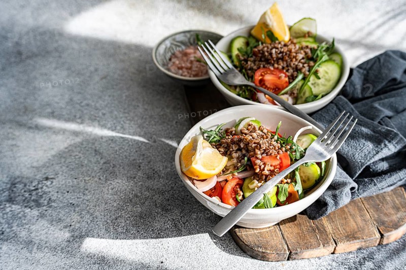 A nutritious lunch featuring fresh rocket, oregano, cherry tomatoes, cucumber, Himalayan salt, red onion, steamed buckwheat, lemon, and organic olive oil, served in bowls