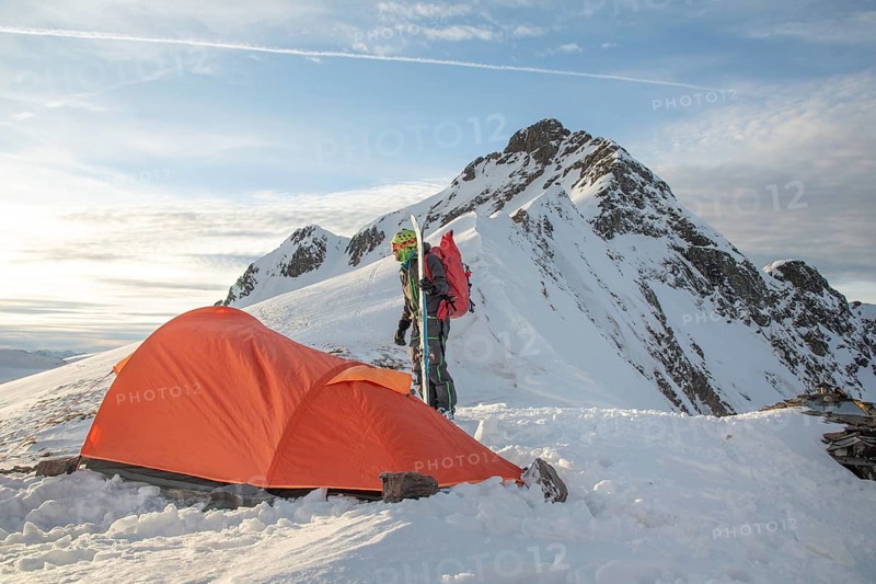 A ski tourer stands near an orange tent on a snowy mountain peak, surrounded by stunning alpine scenery. Perfect depiction of adventure and winter exploration