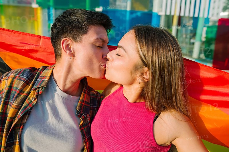 A passionate lesbian couple shares a romantic kiss beneath a flowing rainbow Pride flag. Their loving embrace represents LGBTQIA+ love, unity, and self-expression in a vibrant and meaningful moment