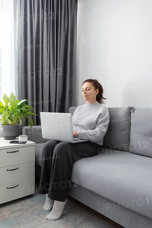 A woman sits on a comfortable gray sofa in a well lit living room, working on a laptop. Nearby is a potted plant and coffee cup on a white side table, under soft curtains
