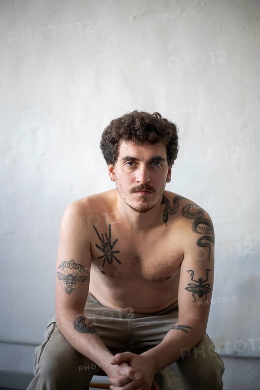 A young man with a short beard and curly hair sits in a relaxed pose against a plain white wall, showcasing intricate tattoos on his arms and chest