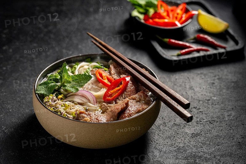 Traditional Vietnamese Beef Pho Noodle Soup in a ceramic bowl, garnished with slices of beef, herbs, red chili, and lime on a dark stone background