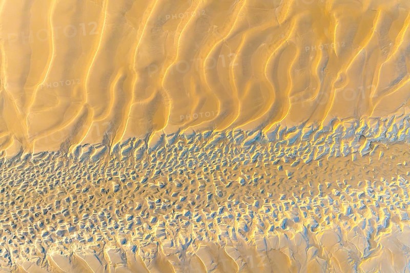 Aerial shot of sand dunes showcasing wavy patterns and textures in a desert landscape. The sun casts soft shadows, highlighting the natural formations and beauty of the terrain